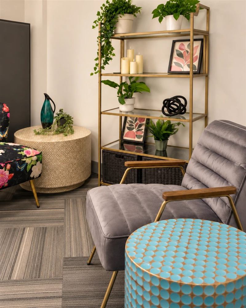 A corner of the therapy room with a floral loveseat, a gold shelf of plants and candles, and soft grey armchairs